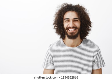 Waist-up shot of friendly happy hispanic bearded guy with curly hair, wearing stylish striped t-shirt, laughing out loud joyfully, smiling broadly from happiness and carefree lifestyle over gray wall