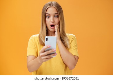 Waist-up shot of shocked and stunned emotive young woman reacting to shocking terrible news reading online from internet gasping opening mouth nervously touchin cheek staring at smartphone screen