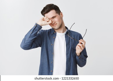 Waist-up Shot Of Tired Male Freelancer Taking Of Glasses As Working Long Near Computer On Gig Economy Project Rubbing Eye And Frowning Being Exhausted Standing Drained And Upset Over Grey Wall