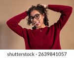 Waist-up studio shot of a young adult female sticking out her tongue at the camera, her hands holding up her hair playfully. She has curly hair and is wearing a red cardigan.