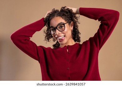 Waist-up studio shot of a young adult female sticking out her tongue at the camera, her hands holding up her hair playfully. She has curly hair and is wearing a red cardigan.