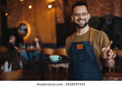 Waiter carrying tray with cappuccino cups in a cafe. Close up of handsome barista holding two cups of coffee in the cafe. Young waiter serving coffee in a cafe and looking at camera. - Powered by Shutterstock