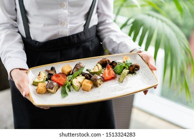 The Waiter Is Holding A Plate Grilled Vegetables With Feta Cheese. Serving On A Plate On A Table. Barbecue Restaurant Menu, A Series Of Photos Of Different Dishes