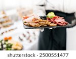 A waiter holding a tray with assorted cold cuts and finger foods at a catered event, with a blurred background of a banquet hall. Perfect for illustrating catering services, hospitality