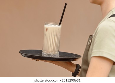 Waiter holding tray with cold, creamy iced coffee, featuring black straw and glass. Server holding chilled iced coffee on black tray, delicious, creamy beverage ideal for hot days, refreshing drink - Powered by Shutterstock