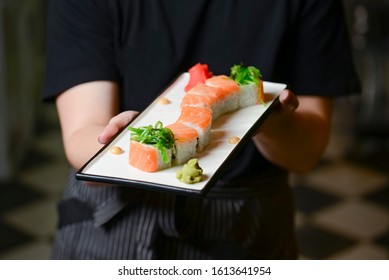 Waiter Holds A Plate Of Tasty Food, Sushi Rolls. Japanese Cuisine In Restaurant. Waiter In A Black Uniform Serving Food.