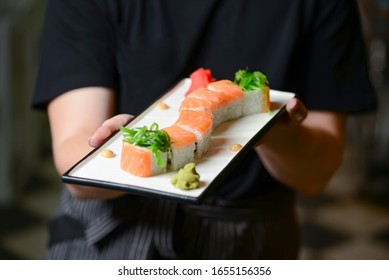 Waiter Holds A Plate Of Tasty Food, Sushi Rolls. Japanese Cuisine In Restaurant. Waiter In A Black Uniform Serving Food In A Restaurant. Waitress At Work.