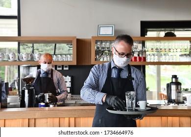 Waiter In A Medical Protective Mask Serves  The Coffee In Restaurant Durin Coronavirus Pandemic Representing New Normal Concept