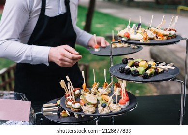 Waiter Places Plates On The Table. Catering Service. Wedding Welcome Food. Fruits On Skewers And Canapes. Welcome Buffet At Wedding Reception.