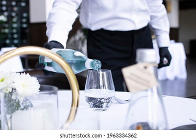 Waiter Pouring Water Into The Glass On The Table, Bottle And Flower Blurred Foreground, In Luxury Fine Dining Restaurant.