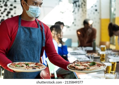 Waiter in protective face mask serving delicious pizza to friends in cozy pizzeria restaurant - Asian waiter with two pizzas Margherita in hands - Original recipe of italian tasty pizza margherita