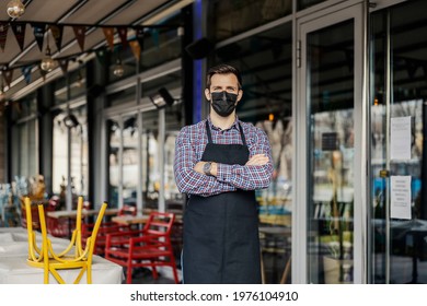 Waiter In A Restaurant. The Server Is Wearing A Plaid Shirt And Apron. He Has His Arms Crossed And Looking Straight At The Camera. Garden Restaurants And Outdoor Cafes. Corona Virus And Face Mask