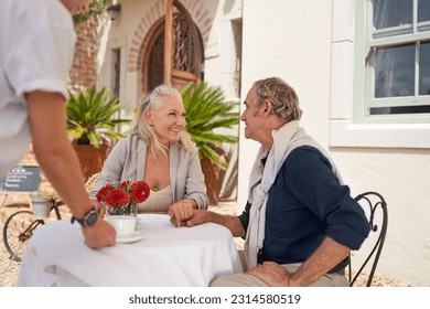 Waiter serving coffee to happy, affectionate retired senior couple holding hands at sidewalk cafe table outside summer villa - Powered by Shutterstock