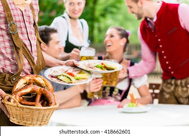 Waiter Serving Food In Bavarian Beer Garden, People Eating And Drinking In Background