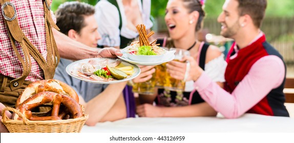 Waiter Serving Food In Bavarian Beer Garden, People Eating And Drinking In Background