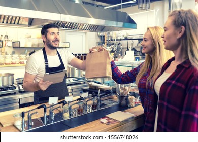 Waiter Serving Takeaway Food To Customers At Counter In Small Family Eatery Restaurant – Smiling Owner Of Trendy Popular Fast Food Delivering To Clients A Online To Go Order In Recycled Paper Bag  