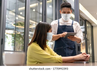 Waiter Wear Protective Face Mask Taking Order From Asian Woman Client With Digital Tablet In Modern Cafe Or Restaurant. People In Public With New Normal Life And Lifestyle