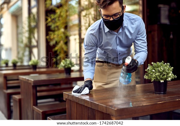 Waiter wearing protective face mask while\
disinfecting tables at outdoor\
cafe.