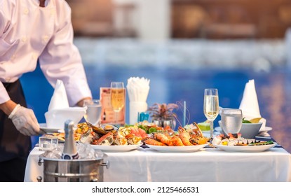 Waiter in white shirt uniform serving dishes of seafood, king pron, crab, champagne, wine on an outdoor table at wedding party or hotel yard garden event. Dinner food catering idea. Selective focus. - Powered by Shutterstock