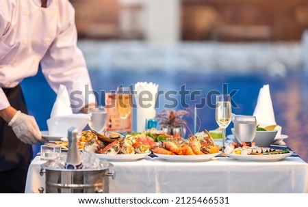 Waiter in white shirt uniform serving dishes of seafood, king pron, crab, champagne, wine on an outdoor table at wedding party or hotel yard garden event. Dinner food catering idea. Selective focus.