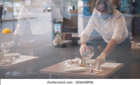 Waiters In Medical Protective Mask Serving Table In Restaurant. View Through Glass Team Of Cafe Employees In Safety Mask And Gloves Setting Table Preparing For Reopening