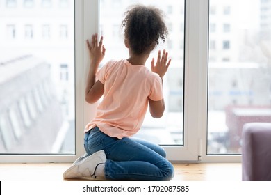 Waiting african adolescent girl sitting alone on windowsill in living room at home. Young single female person thinking staring at window. Sad daughter feel upset. Youth problems, adoption concept