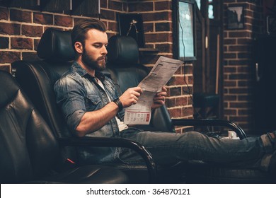 Waiting for appointment. Handsome young bearded man reading newspaper while sitting in comfortable chair at barbershop - Powered by Shutterstock