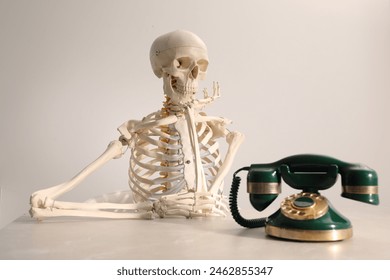 Waiting concept. Human skeleton at table with corded telephone against light grey background