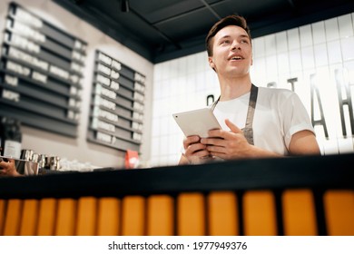 Waiting For Customers After The Pandemic Sells Coffee With A Takeaway. The Cafe Opened. Records Business Expenses In A Tablet In A Table. A Male Employee In A Restaurant Uniform Is Working.