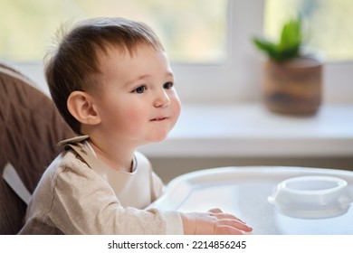 Waiting For Food, Baby Todd Is Sitting At An Empty Table. A Child In Anticipation Of Lunch On A High Children Chair. Kid Aged One Year And Two Months