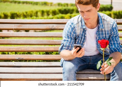 Waiting For His Girlfriend. Worried Young Man Holding Single Rose And Looking At His Mobile Phone While Sitting On The Bench