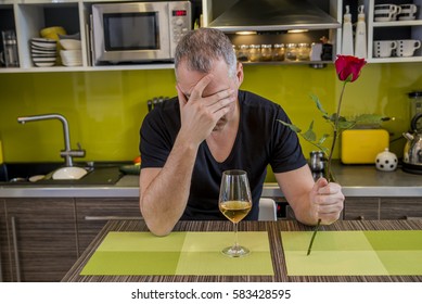 Waiting For His Girlfriend. Worried Young Man Holding Single Rose And Looking Depressed While Sitting In The Kitchen