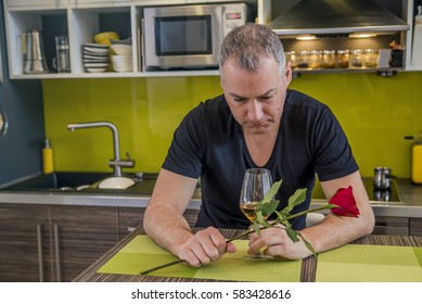 Waiting For His Girlfriend. Worried Young Man Holding Single Rose And Looking Depressed While Sitting In The Kitchen