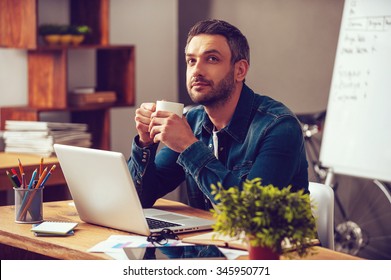Waiting for inspiration. Thoughtful young man holding coffee cup and looking away while sitting at his working place in office
