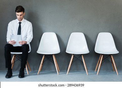 Waiting for interview. Confident young businessman holding paper while sitting on chair against grey background
