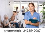 In waiting room, friendly young female medical officer, clinic nurse in surgical suit, keeps list of patients and waits for patients to arrive at outpatient department