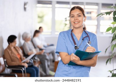 In waiting room, friendly young female medical officer, clinic nurse in surgical suit, keeps list of patients and waits for patients to arrive at outpatient department - Powered by Shutterstock