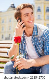 I Am Waiting For You! Happy Young Man Holding Single Rose And Talking On The Mobile Phone While Sitting On The Bench