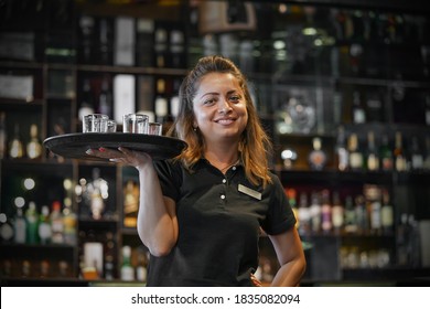 Waitress Carries A Whiskey Glass On A Tray In Hotel Restaurant, Bar. The Concept Of Service. Shelves With Bottles Of Alcohol In The Background. Night Time.