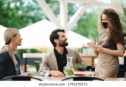 Waitress With Face Mask Serving Happy Couple Outdoors On Terrace Restaurant.