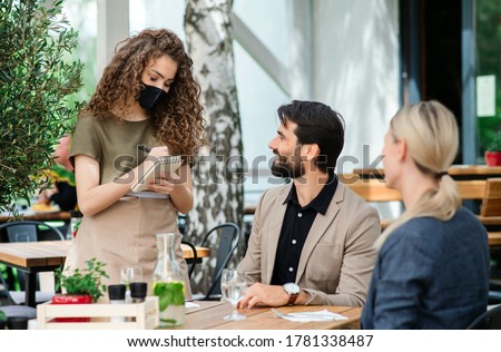 Waitress with face mask serving happy couple outdoors on terrace restaurant.
