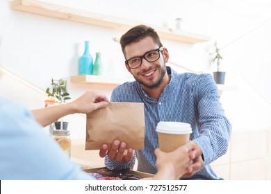 Waitress Giving Coffee To Go And Paper Bag With Take Away Food To Smiling Young Client In Cafe