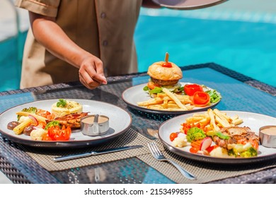 Waitress Hand Serving Lunch In Restaurant At Luxury Hotel. Asian Woman Waiter Put Delicious Food Plates On Table In Outdoors Pool Cafe In Tropical Resort. Summer Travel, Holidays, Vacations Concept.