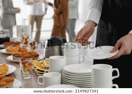 Waitress near table with dishware and different delicious snacks during coffee break, closeup
