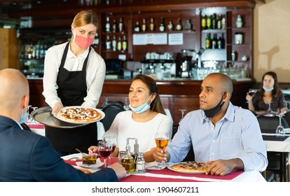 Waitress in protective mask serving delicious pizza to friends in cozy restaurant
