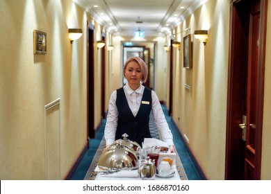 Waitress In Uniform Walking Along The Hall While Delivering Tray With Food In A Room Of Hotel. Room Service. Horizontal Shot. Front View