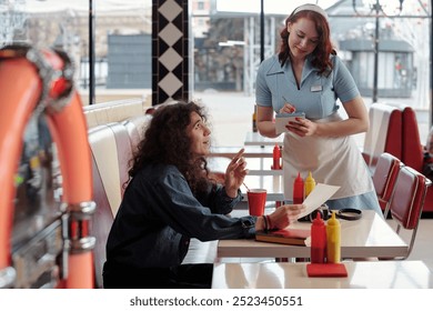 Waitress in vintage uniform taking order from customer sitting in classic American diner booth checking menu while holding notepad and pen - Powered by Shutterstock