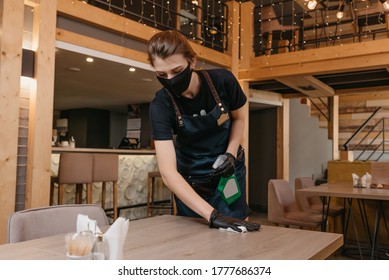 A Waitress Who Wears A Black Medical Face Mask And Disposable Medical Gloves Is Holding A Bottle With Sanitizer And Cleaning A Table With A Rag In A Restaurant. A Barista Is Waiting For Clients.