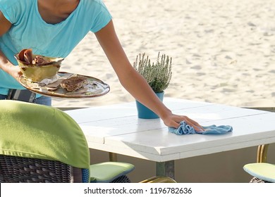 The Waitress Wipes The Table In A Cafe On The Beach. A Waitress Is Holding A Tray With Dirty Dishes And Leftover Food. Waitress Cleaning The Table In A Restaurant. The Concept Of Service.