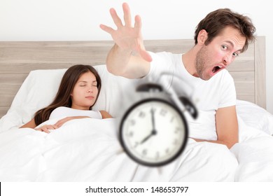 Wake Up - Couple Waking Up Early Throwing Alarm Clock. Funny Bed Concept With Young Interracial Couple Waking Up Late. Man Throwing Alarm Clock, And Woman Sleeping. Asian Female, Caucasian Male Models